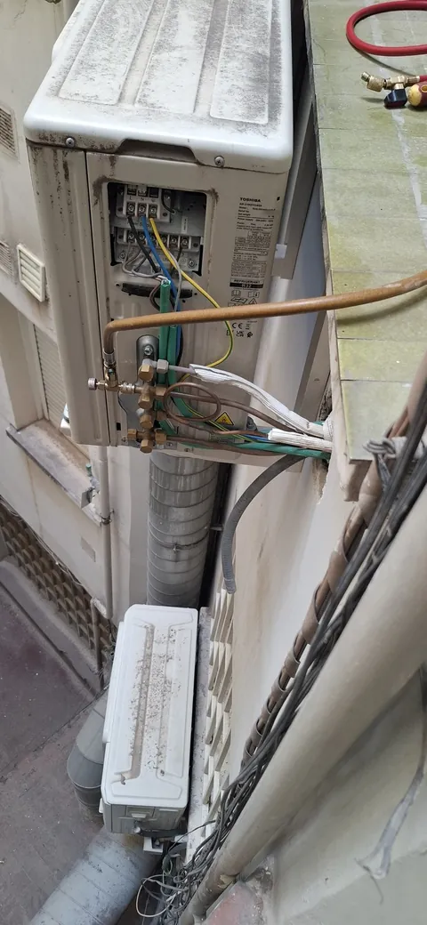 Unidad exterior de aire acondicionado Toshiba en una fachada, con la tapa de bornas abierta y tuberías de cobre con válvulas