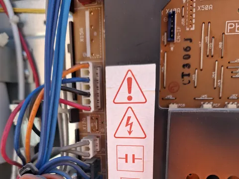 Cuadro eléctrico abierto de una unidad exterior: placa electrónica marrón, conectores blancos con cables azules, rojos y naranjas y una pegatina de aviso de riesgo eléctrico